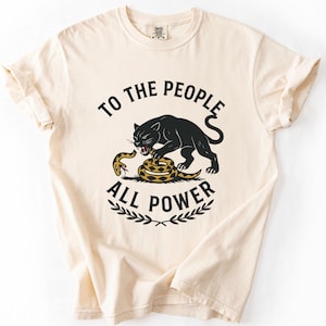 Può includere: Una t-shirt a maniche corte color crema con la grafica di una pantera nera che attacca un serpente giallo e marrone. Il testo "TO THE PEOPLE" è arcuato sopra la pantera e "ALL POWER" sotto, con un piccolo disegno di foglie alla base.