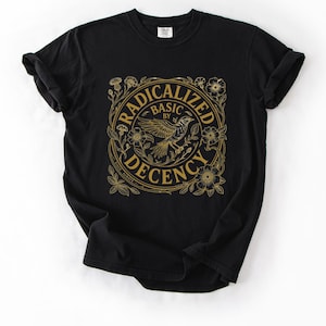 Op de afbeelding: Zwart T-shirt met een gouden ontwerp. Het ontwerp toont een vogel, bloemen en de woorden "RADICALIZED BY BASIC DECENCY" in een cirkelvormige lijst. Het shirt is gemaakt van een zacht materiaal.