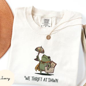 May include: An ivory t-shirt featuring a graphic of a green frog wearing a patterned cape, holding a stained-glass style lamp, and carrying a tote bag with a framed picture inside. The text below the frog reads WE THRIFT AT DAWN.