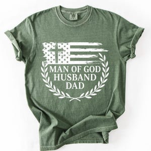 Könnte beinhalten: Ein salbeigrünes Kurzarm-T-Shirt mit einem weißen Grafikdruck einer abgenutzten amerikanischen Flagge mit einem Kreuz im oberen linken Feld. Unter der Flagge steht der Text MAN OF GOD HUSBAND DAD, umrahmt von einem weißen Lorbeerkranz.