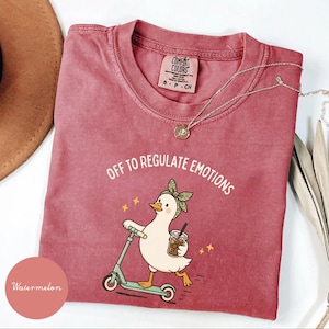 Puede incluir: Una camiseta color rosa polvoriento con el gráfico de un pato blanco con una diadema verde de lunares, montado en un patinete verde claro y sosteniendo un café helado. El texto dice OFF TO REGULATE EMOTIONS. La camiseta tiene una etiqueta Comfort Colors.
