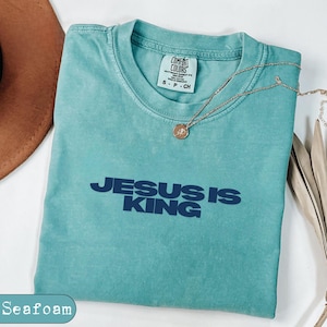 Puede incluir: Una camiseta de manga corta en color verde espuma de mar con cuello redondo. La prenda presenta el texto "JESUS IS KING" impreso en letras azul oscuro en el pecho. Un pequeño collar dorado con un colgante circular descansa sobre el cuello.
