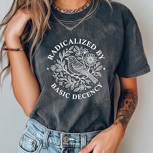 Pode incluir: Camiseta cinza escuro com o texto "RADICALIZED BY BASIC DECENCY" em um design circular branco com um pássaro e elementos florais. A camiseta tem mangas curtas e gola redonda.