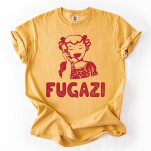 May include: Mustard yellow t-shirt with a red graphic of a smiling person holding a telephone receiver. The word "FUGAZI" is printed in red block letters below the graphic. The shirt has rolled-up sleeves.