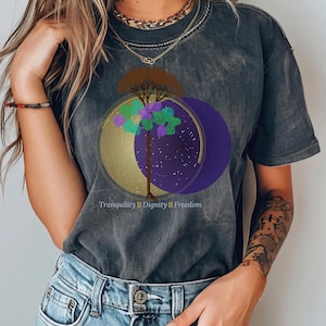 Può includere: Una t-shirt grigio antracite con effetto lavaggio acido che presenta la grafica di un albero con fogliame verde e viola su cerchi sovrapposti dorati e viola scuro con un motivo a stelle. Il testo "Tranquility | Dignity | Freedom" è stampato sotto.