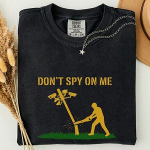 Może przedstawiać: Czarny t-shirt z okrągłym dekoltem z żółtym napisem DON'T SPY ON ME nad grafiką przedstawiającą osobę używającą piły łańcuchowej do ścięcia słupa energetycznego z kamerami bezpieczeństwa. Koszulka posiada metkę marki Comfort Colors przy szyi.