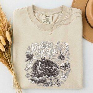 May include: A beige t-shirt featuring a dark brown graphic print with the text Midnight Reader. The illustration depicts a person reading a book with a cat, a crescent moon, a skull, a scroll, a quill, and stars. The shirt has a crew neckline.