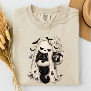 Könnte beinhalten: Ein beiges T-Shirt mit Rundhalsausschnitt und einer Grafik eines Geistes, der in ein Laken gehüllt ist und eine schwarze Tasse mit der Aufschrift MEH und einem kleinen Geistersymbol hält. Der Geist hält eine flauschige schwarze Katze.
