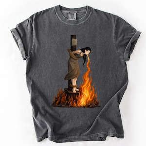 Puede incluir: Camiseta gris oscuro con un gráfico de una mujer atada a una estaca, con llamas que se elevan desde abajo. La mujer tiene el pelo largo y negro y lleva un vestido marrón, con llamas naranjas saliendo de su boca.
