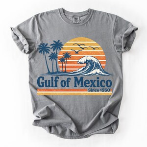 Puede incluir: Una camiseta gris con un gráfico de una puesta de sol, palmeras y una gran ola oceánica. El texto dice Gulf of Mexico Since 1550. El diseño utiliza colores naranja, amarillo y azul con una textura desgastada y envejecida en el estampado.