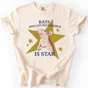 Puede incluir: Camiseta color crema con un gráfico que presenta una rata dentro de una estrella verde, rodeada de estrellas más pequeñas. El texto dice "RATS SPELLED BACKWARDS IS STAR" en azul marino. La camiseta tiene mangas cortas y cuello redondo.