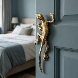 Puede incluir: Manija de puerta dorada con forma de lagarto montada en una puerta azul. El mango está detallado con escamas y garras texturizadas, agregando un elemento decorativo único. La puerta es parte de un dormitorio, con una cama visible en el fondo.