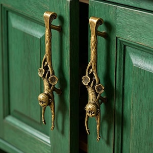 Peut inclure: Deux poignées de placard en métal couleur laiton antique en forme de singes suspendus à des branches. Chaque poignée présente une figurine de singe détaillée avec une fourrure texturée et des fleurs, fixée verticalement sur des portes en bois vert.