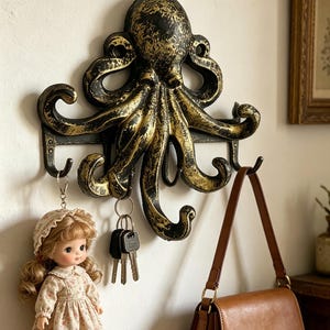 Pode incluir: Um porta-chaves decorativo de polvo montado na parede em preto e dourado. O design do polvo tem vários ganchos para chaves e uma bolsa. Uma boneca e uma bolsa marrom penduram-se nos ganchos.