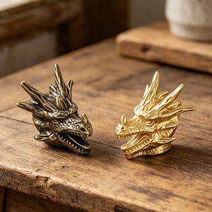 以下が含まれることがあります： 木製の表面に置かれた、ブロンズとゴールドの2つのドラゴンの頭の彫刻。それぞれに詳細な鱗、角、開いた口があります。彫刻は、ファンタジーや神話のテーマの装飾品である可能性があります。