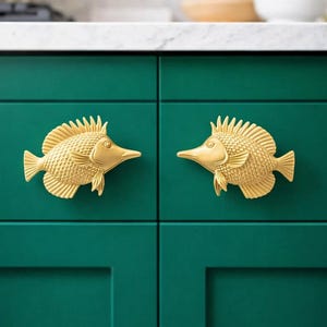 May include: Two gold-coloured fish-shaped cabinet pulls on a dark teal cabinet. The cabinet is part of a kitchen setup, with a white countertop and a blurred background.