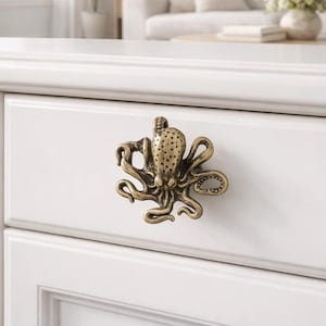 Pode incluir: Puxador de armário em forma de polvo de bronze em uma gaveta branca. O polvo tem tentáculos detalhados e uma cabeça texturizada. A gaveta faz parte de um armário branco, com um fundo desfocado de uma sala de estar.