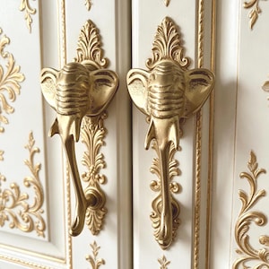 Peut inclure: Gros plan sur deux poignées de porte d'armoire dorées en forme d'éléphant. Les poignées sont fixées à des portes d'armoires blanches avec des bordures décoratives et des motifs floraux dorés. Les poignées d'éléphant sont détaillées avec une peau et une trompe texturées.