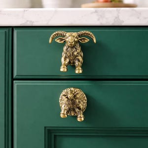 May include: Two brass cabinet pulls in the shape of a ram and a sheep are mounted on a dark green cabinet. The ram pull is above the sheep pull. The pulls have detailed features and a vintage aesthetic.