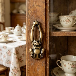 May include: A brass monkey-shaped cabinet pull is mounted on a distressed wooden cabinet door. The monkey is hanging from a bar, with its body and face detailed. The cabinet door is part of a display cabinet with floral teacups and saucers visible inside.
