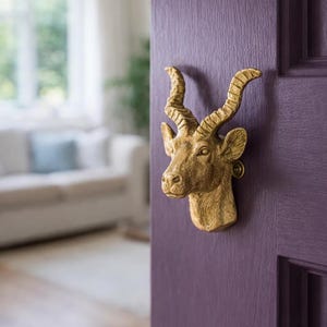 May include: A gold-colored door knocker in the shape of a deer head is mounted on a purple door. The deer head has detailed antlers and facial features. The door is a solid color with a textured surface.