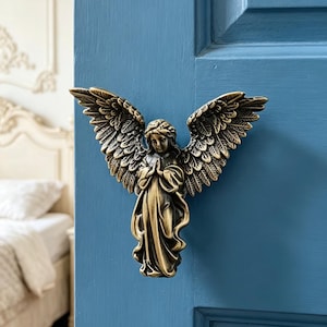 May include: A bronze-colored angel door knocker with outstretched wings, mounted on a blue door. The angel is depicted in a praying pose, with detailed feathering on the wings. The background includes a glimpse of a white bed and wall trim.