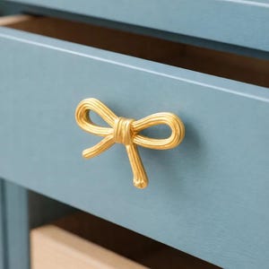 Puede incluir: Un tirador dorado en forma de lazo está montado en un cajón azul claro. El cajón está parcialmente abierto, revelando un interior de madera. El tirador en forma de lazo añade un toque decorativo al mueble.