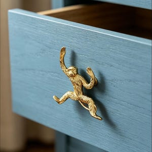 K&ouml;nnte beinhalten: Ein goldfarbener Schubladengriff aus Metall in Form eines Affen, der an einer blau lackierten Holzschubladenfront befestigt ist. Die Affenfigur ist in einer springenden Pose mit texturierten Fell-Details dargestellt und dient als Griff.