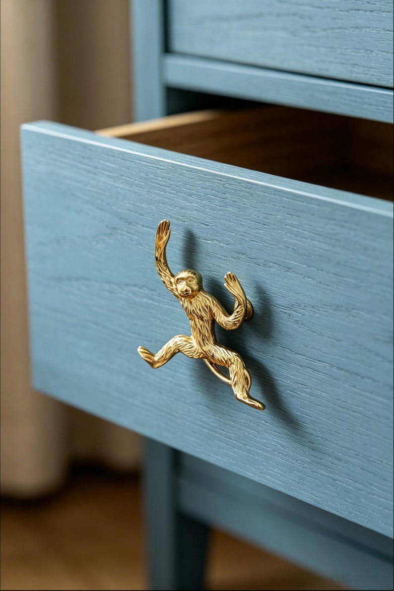 K&ouml;nnte beinhalten: Ein goldfarbener Schubladengriff aus Metall in Form eines Affen, der an einer blau lackierten Holzschubladenfront befestigt ist. Die Affenfigur ist in einer springenden Pose mit texturierten Fell-Details dargestellt und dient als Griff.