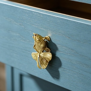 May include: A metallic gold-toned drawer pull shaped like a goldfish with detailed scales and flowing fins, mounted on a blue-painted wooden drawer front with a visible wood grain texture and an open top edge showing the interior construction.
