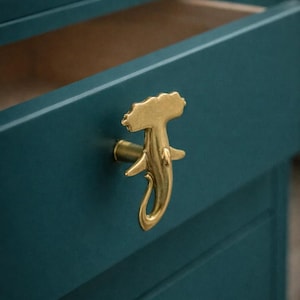 May include: A close-up of a teal-colored drawer with a gold-colored, stylized handle. The handle has a unique, organic shape, possibly resembling a sea creature. The drawer is partially open, revealing a wooden interior.