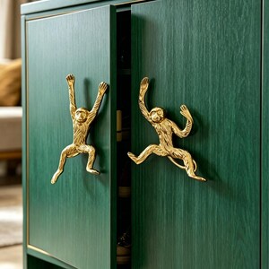 K&ouml;nnte beinhalten: Ein dunkelgr&uuml;ner Holzschrank mit zwei T&uuml;ren und goldfarbenen, affenf&ouml;rmigen Metallgriffen. Der Schrank weist eine sichtbare Holzmaserung und eine d&uuml;nne goldene Verzierung an den T&uuml;rkanten auf. Die Affengriffe sind in dynamischen Posen gestaltet.