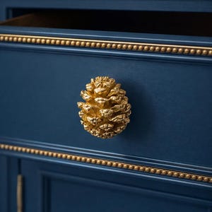 Puede incluir: Primer plano de un armario azul marino con detalles dorados. Un pomo dorado en forma de piña se centra en la puerta del armario. El armario tiene adornos dorados con pequeños detalles redondos. El conjunto es elegante y decorativo.