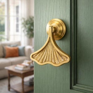 Puede incluir: Un tirador de puerta dorado con un diseño en forma de abanico, montado en una puerta verde. El tirador tiene una superficie detallada y texturizada y una base circular. La puerta está pintada de color verde apagado.