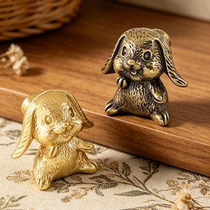 Può includere: Due piccole e dettagliate statuette di coniglietti, in tonalità oro e bronzo. I coniglietti sono seduti con lunghe orecchie e facce sorridenti. Le statuette sono posizionate su una superficie di legno, suggerendo un oggetto decorativo o da collezione.