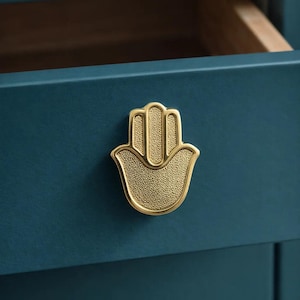 May include: A gold-colored Hamsa hand-shaped drawer pull with a textured surface is mounted on a teal blue drawer. The drawer is partially open, revealing a wooden interior. The hardware adds a decorative touch.