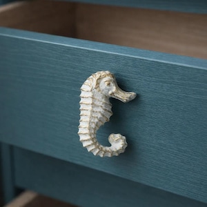 Può includere: Pomello per mobile a forma di cavalluccio marino con finitura bianca anticata montato su un cassetto blu teal. La maniglia a forma di cavalluccio marino aggiunge un tema costiero o nautico ai mobili. Il cassetto è aperto, rivelando un interno in legno chiaro.