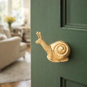 May include: A gold-colored snail-shaped door handle mounted on a dark green door. The snail's body is textured, and the handle is in a whimsical design. The door is part of a room with a blurred background.