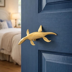 Può includere: Maniglia dorata a forma di plesiosauro, montata su una porta blu scuro. La maniglia è una scultura dettagliata del rettile marino preistorico. La porta fa parte di un interno di camera da letto.