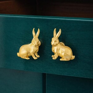 May include: Two gold-colored bunny-shaped cabinet pulls mounted on a teal-colored cabinet door. The bunny pulls are detailed with upright ears and seated poses. The cabinet has a recessed panel design.
