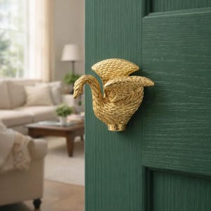Op de afbeelding: Een goudkleurige zwaanvormige deurklink gemonteerd op een donkergroene deur. De zwaan heeft gedetailleerde veertjesstructuren en een gebogen nek. De deur maakt deel uit van een kamer met een vage achtergrond.