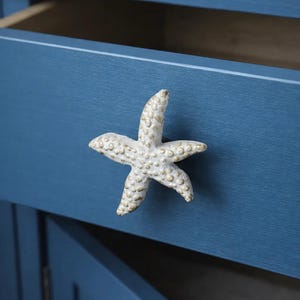 Op de afbeelding: Een witte, stervormige kastknop met gouden accenten is bevestigd aan een blauwe kastdeur. De kastdeur is open en onthult een houten interieur. De knop voegt een kust- of nautisch thema toe aan het meubel.