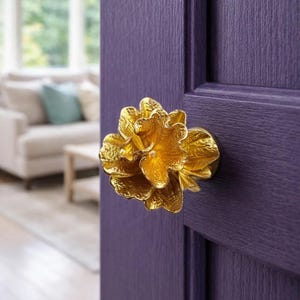 Könnte beinhalten: Ein goldfarbener, blütenförmiger Türknauf ist an einer strukturierten, lila Tür angebracht. Die Tür ist teilweise geöffnet und gibt den Blick auf ein Wohnzimmer mit Sofa und Fenster frei.