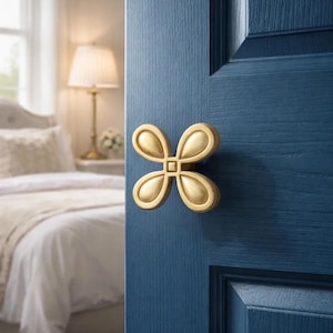 Op de afbeelding: Een close-up van een blauwe deur met een goudkleurige, vierbladige bloemvormige deurklink. De deur maakt deel uit van een slaapkamer met een bed en een lamp op de achtergrond. De deurklink is het middelpunt.
