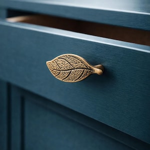 Puede incluir: Tirador de cajón en forma de hoja de latón montado en un mueble azul oscuro. El cajón está parcialmente abierto, revelando un interior de madera. El diseño de la hoja presenta intrincados detalles de veteado, añadiendo un toque de elegancia inspirada en la naturaleza.