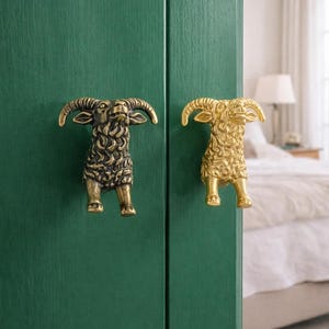 May include: Two ram-shaped cabinet pulls. One is bronze, the other gold. They are mounted on a dark green cabinet door. The pulls have detailed horns and textured fur. The cabinet is in a bedroom.