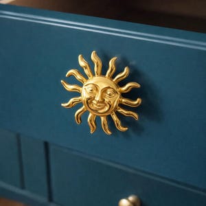 May include: A close-up of a dark teal cabinet with a gold sun-shaped drawer pull. The sun has a smiling face and radiating rays. A smaller gold knob is visible below the sun pull.