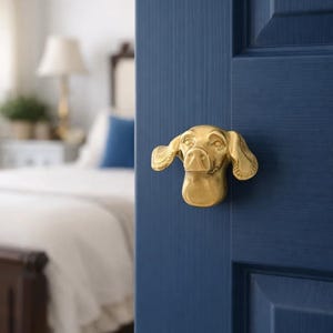 Könnte beinhalten: Ein messingfarbener Türgriff in Hundeform ist an einer marineblauen Tür angebracht. Der Kopf des Hundes ist detailliert mit sichtbaren Ohren, Nase und Augen. Der Hintergrund zeigt ein verschwommenes Schlafzimmer mit Bett und Lampe.