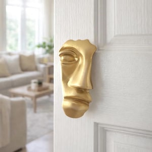 Puede incluir: Un aldaba dorada con forma de rostro humano está montada en una puerta blanca. La cara tiene un diseño estilizado, ligeramente abstracto, con los ojos cerrados y una nariz prominente.