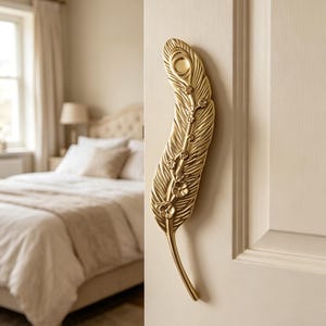 May include: A gold-colored, feather-shaped door handle with intricate detailing. The handle is mounted on a cream-colored door and features small floral accents along its stem. The design evokes a sense of elegance and sophistication, suitable for interior decor.